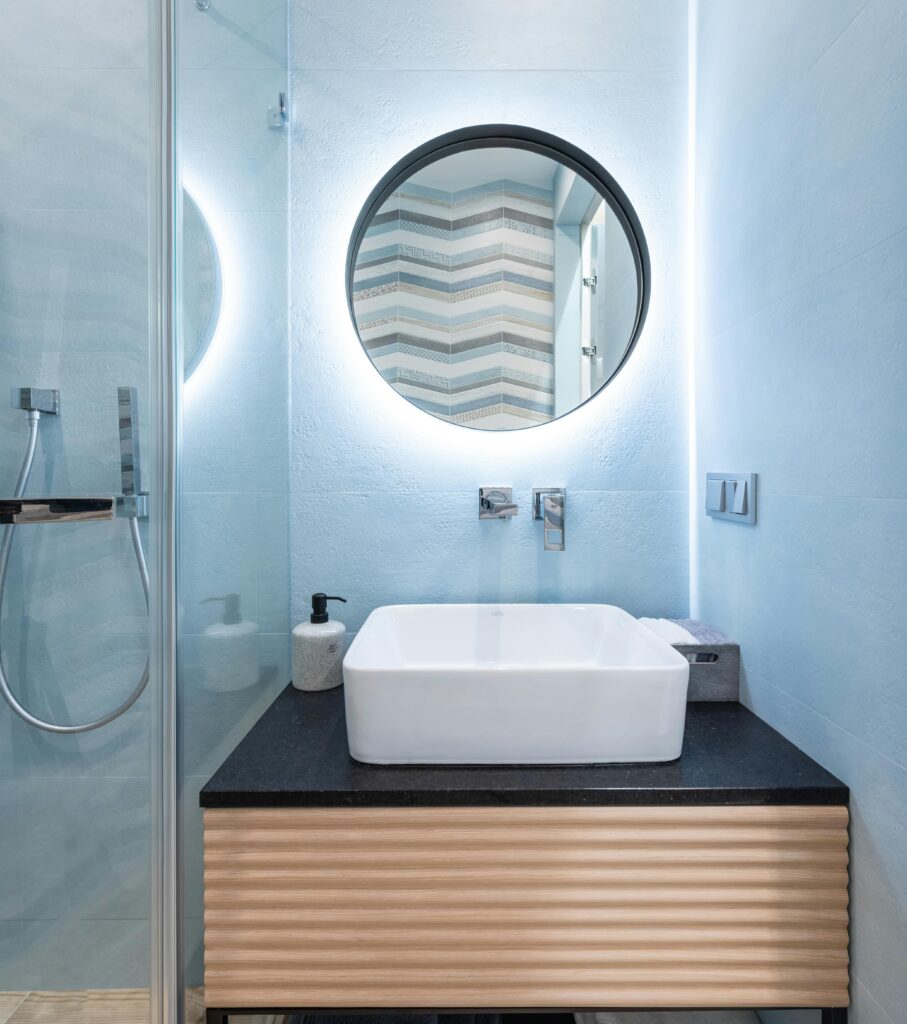 Interior of modern bright restroom with sink and tap on counter under round mirror on wall next to glass wall to shower