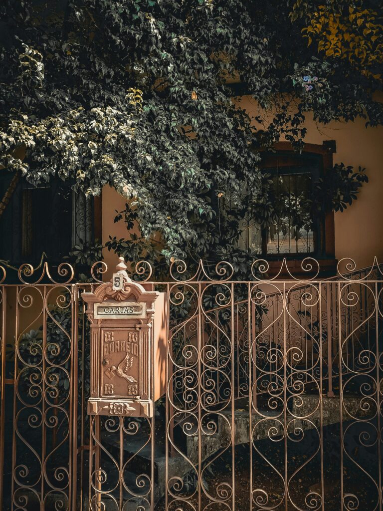 Ornate iron gate with mailbox, vintage design surrounded by lush foliage.