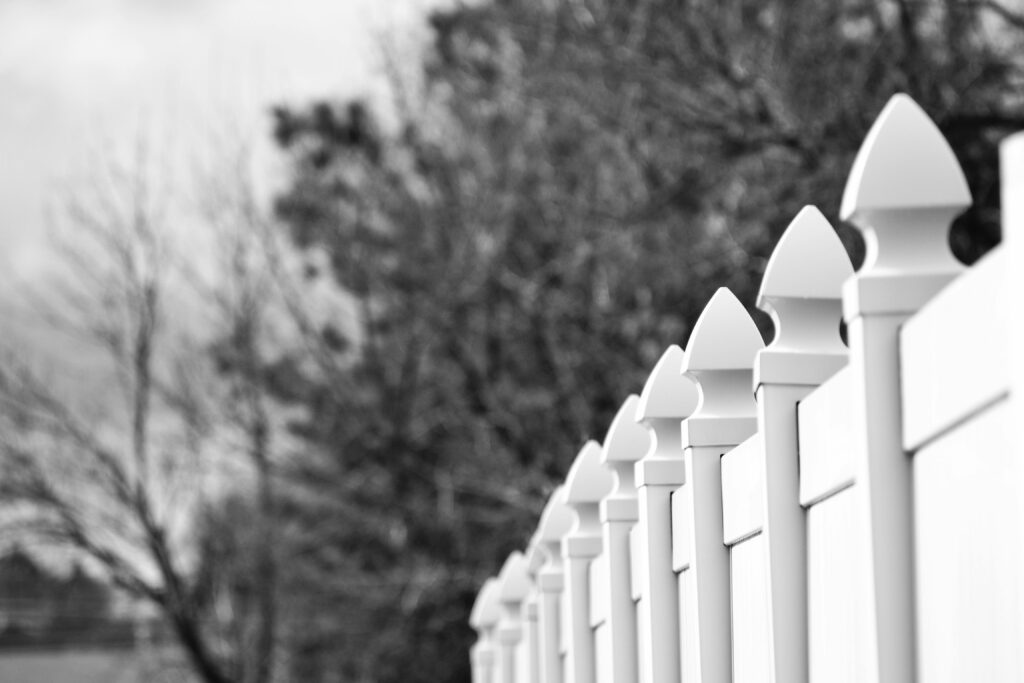 Black and white image of a row of decorative fence posts with blurred winter trees in the background.