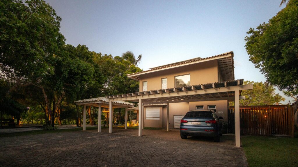 A modern tropical house with a pergola and driveway at twilight, surrounded by lush greenery.